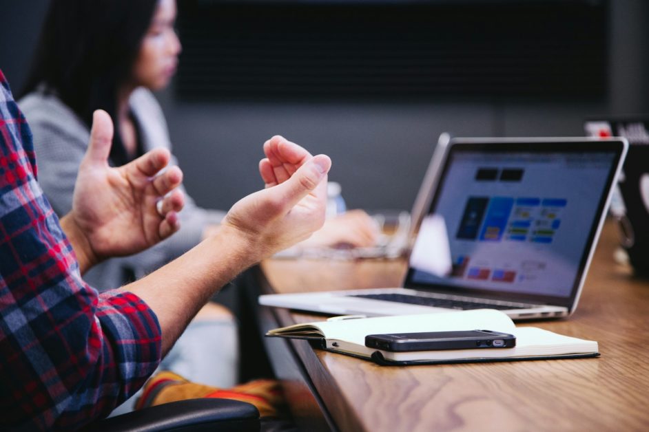 Close up of hands of individual during a meeting. On the table is a laptop. In the background is a colleague.